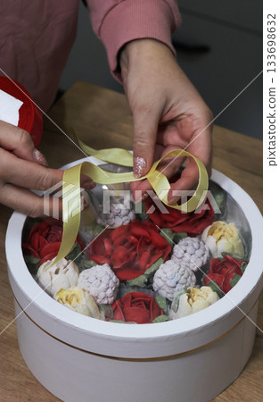 Female entrepreneur assembling marshmallow flowers in a hat box for a gift, tying it with colorful ribbons, using scissors, marshmallow tulips and roses. Oblique Angle. Female entrepreneur assembling marshmallow flowers in a hat box for a gift, tying it with colorful ribbons, using scissors, marshmallow tulips and roses. Oblique Angle. 133698632