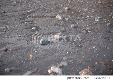 Smooth pebbles on a sandy shoreline, partially submerged in shallow water. The central pebble is distinctly larger, rounded, and grey, surrounded by smaller stones of various colors including muted re 133698685