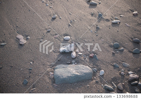 Smooth pebbles on a sandy shoreline, partially submerged in shallow water. The central pebble is distinctly larger, rounded, and grey, surrounded by smaller stones of various colors including muted re 133698688