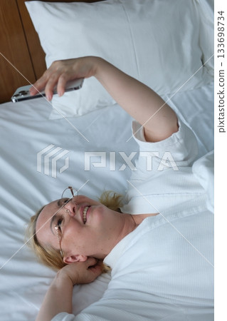 Cheerful adult woman taking selfie with phone while resting on bed in the morning. Happy millennial female sharing self portrait on social media network. 133698734