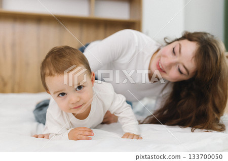Cozy moment of a young woman and baby relaxing together at home, lying on a soft bed and smiling in natural light. 133700050