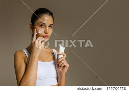 Young woman applying face cream while holding a cosmetic tube, demonstrating a hydrating skincare step on a clean studio background. 133700758