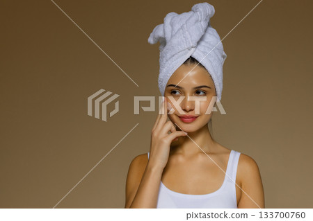 Young woman applying skincare cream to her cheek after a shower, wearing a towel on her head and showing a gentle beauty routine. Young woman applying skincare cream to her cheek after a shower, wearing a towel on her head and showing a gentle beauty routine. 133700760