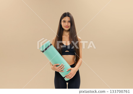 Young brunette woman in black sportswear holds a turquoise yoga mat, posing against a beige background, fitness and healthy lifestyle concept. 133700761