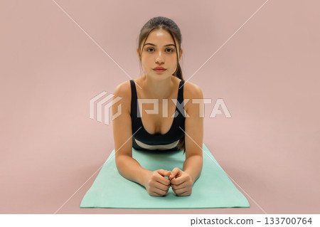 Young woman lying on a yoga mat during a workout, looking forward while practicing a fitness plank variation on a pastel background Young woman lying on a yoga mat during a workout, looking forward while practicing a fitness plank variation on a pastel background 133700764