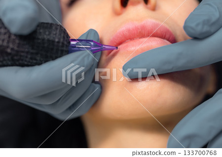 Close up of a cosmetologist performing permanent lip makeup, tattooing natural lip color enhancement with sterile tools in a professional beauty studio. 133700768