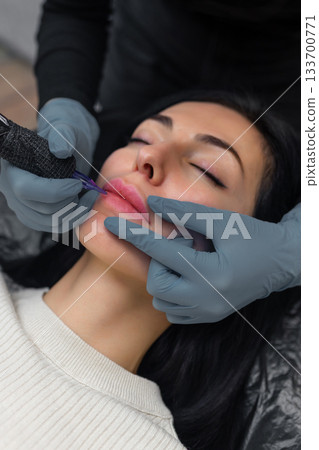Close up of a cosmetologist performing permanent lip makeup, tattooing natural lip color enhancement with sterile tools in a professional beauty studio. Close up of a cosmetologist performing permanent lip makeup, tattooing natural lip color enhancement with sterile tools in a professional beauty studio. 133700771