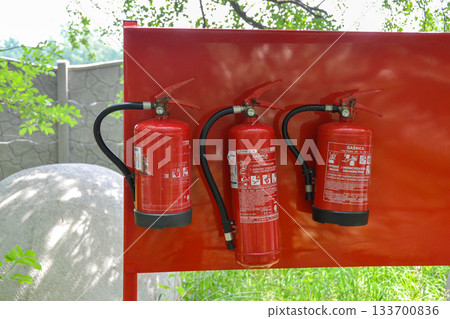 Three fire extinguishers are mounted on a crimson wall, ready to combat any unexpected flames. Each cylinder contains lifesaving gas for emergency situations 133700836