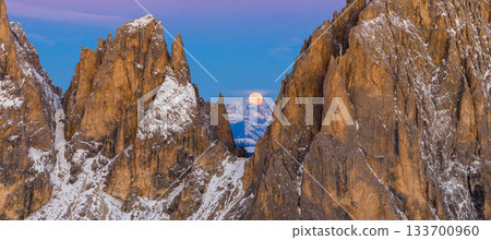 Panoramic view of Dolomite mountain peaks under a glowing full moon. Early winter landscape with snow-covered summits in soft dawn light. Captured in high resolution, ideal for large prints. 133700960