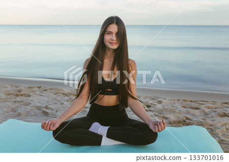 Woman meditating on yoga mat by the sea, yoga meditation, wellness routine, mindfulness practice, beach fitness, calm breathing, relaxing seaside yoga session at sunrise. 133701016