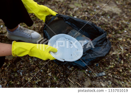 Volunteer collecting plastic waste and litter into a black trash bag during beach cleanup, highlighting pollution, environmental protection, sustainability, and eco awareness. 133701019