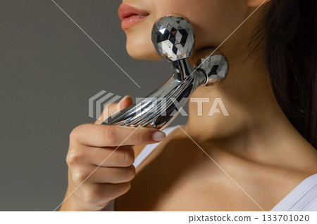 Close-up of a young woman massaging her neck with a metal 3D roller, demonstrating a tightening and relaxing skincare routine at home. 133701020