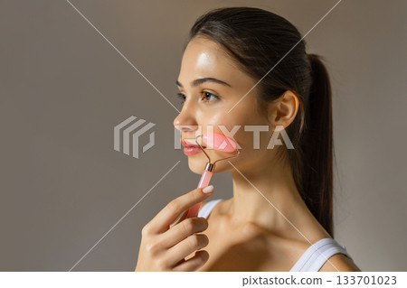 Young woman using a pink facial roller on her cheek, demonstrating a relaxing skincare massage routine on a soft studio background. Young woman using a pink facial roller on her cheek, demonstrating a relaxing skincare massage routine on a soft studio background. 133701023