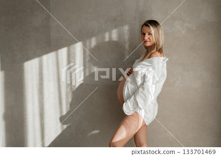 34 week, A pregnant woman stands in soft sunlight wearing an oversized white shirt. Gentle shadows on the textured wall create a natural, warm atmosphere ideal for lifestyle, maternity, wellness and 133701469