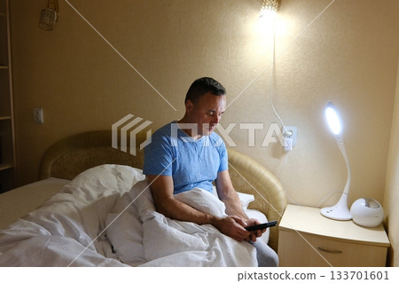 An adult male man sitting on a bed at night, smartphone screen, deep in thought. dim lighting and focused expression. Themes of insomnia, social media addiction, stress, late-night scrolling 133701601