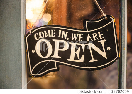Closeup of a vintage open sign hanging on a shop window with warm bokeh lights in the background. 133702009