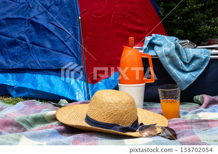 Enjoying summer leisure in the countryside. A colorful tent pitched as a comfortable base for outdoor relaxation. With a picnic blanket spread. Straw hat protecting from the sun 133702054