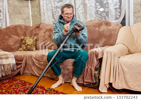 Cleaning vacuum brush, middle aged man focuses on removing dirt from household tool, facial expression shows clear disgust during simple task. Cleaning vacuum brush, middle aged man focuses on removing dirt from household tool, facial expression shows clear disgust during simple task. 133702243
