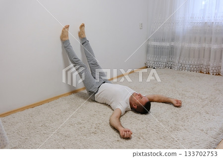 A grown man lies on the floor on a carpet at home with his legs raised up on the wall and his arms spread out to the sides. 133702753