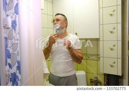 A man shaves his face in a bathroom with butterflies on the wall and light green tiles. white man shaving with brush and shaving foam looking in the mirror A man shaves his face in a bathroom with butterflies on the wall and light green tiles. white man shaving with brush and shaving foam looking in the mirror 133702775