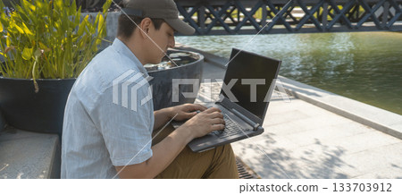 A man is sitting in a park and working on a laptop outdoors. A man is sitting in a park and working on a laptop outdoors. 133703912
