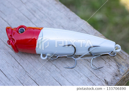 Eye-catching artificial lure in white and red on a rustic wooden board. Fishing and outdoor recreation concept. River sports. Fishing enthusiast gear. 133706890