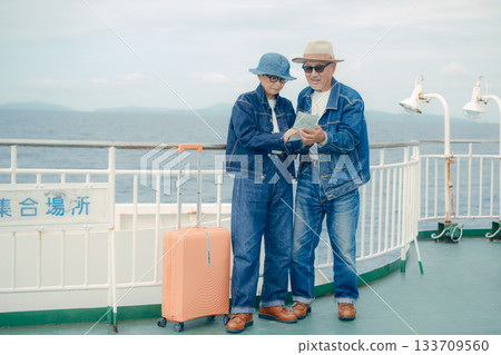 Senior couple with suitcases looking at a smartphone and pamphlet on the ferry deck 133709560
