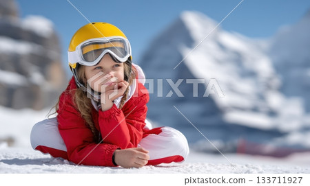 Five-year-old girl in a red-white ski suit and yellow helmet sits thoughtfully on a snow-covered mountain, enjoying the winter landscape 133711927