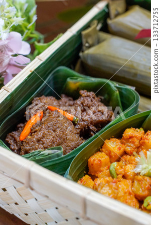 Indonesian Rendang Beef and Spicy Potato Balado Served in Traditional Banana Leaf Containers Inside a Woven Bamboo Basket, Authentic Southeast Asian Cuisine Close Up with Rich Textures and Vibrant Col 133712755