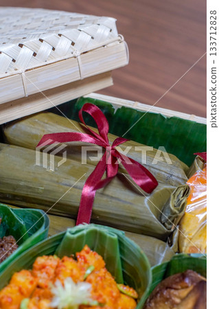Traditional Indonesian lontong wrapped in banana leaves with red ribbon inside a woven bamboo hamper, close-up of authentic Southeast Asian cuisine and eco-friendly food packaging Traditional Indonesian lontong wrapped in banana leaves with red ribbon inside a woven bamboo hamper, close-up of authentic Southeast Asian cuisine and eco-friendly food packaging 133712828