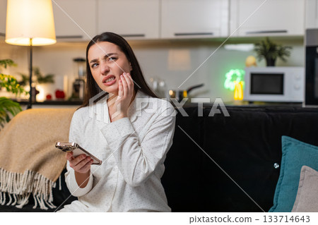 Young woman holds cheek tooth pain searches dentist on smartphone books visit dental care at night Young woman holds cheek tooth pain searches dentist on smartphone books visit dental care at night 133714643