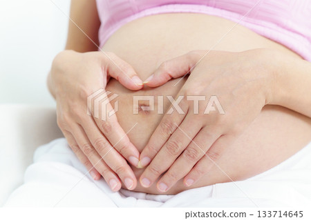 Asian pregnant woman relaxing at home. Asian pregnant woman is sitting and relaxing on a sofa in living room and putting her hands on stomach in the heart shape. 133714645