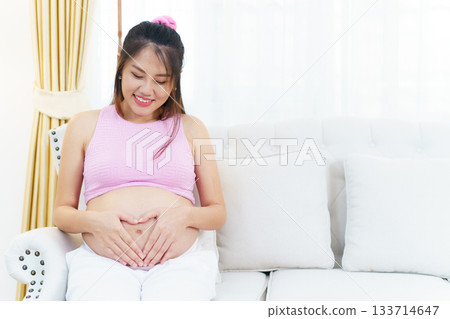 Asian pregnant woman relaxing at home. Asian pregnant woman is sitting and relaxing on a sofa in living room and putting her hands on stomach in the heart shape. 133714647