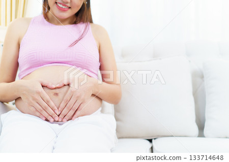 Asian pregnant woman relaxing at home. Asian pregnant woman is sitting and relaxing on a sofa in living room and putting her hands on stomach in the heart shape. 133714648