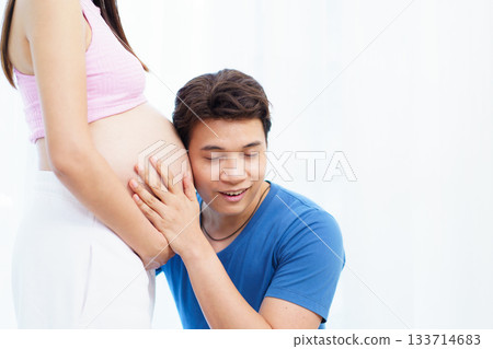 A man is pressing his ear against his pregnant wife's belly while gently embracing it to listen to the sounds and movements of their baby. He is filled with joy as he connects with their unborn child. A man is pressing his ear against his pregnant wife's belly while gently embracing it to listen to the sounds and movements of their baby. He is filled with joy as he connects with their unborn child. 133714683