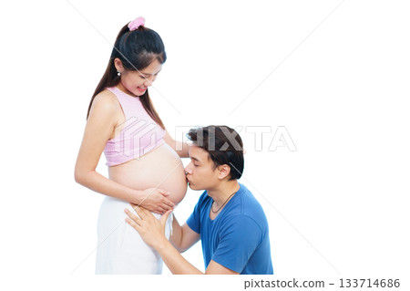 A man is pressing his ear against his pregnant wife's belly while gently embracing it to listen to the sounds and movements of their baby. He is filled with joy as he connects with their unborn child. 133714686