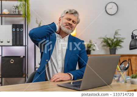 Middle-aged businessman grimaces sudden neck pain clutching neck seeking relief during work session 133714720