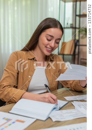 Young woman reviewing receipts nodding satisfied after accurate calculations writing notes at desk 133714732