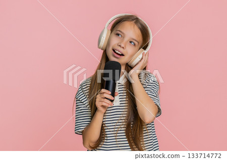 Child girl wearing wireless headphones sings into microphone performing imaginary concert happily Child girl wearing wireless headphones sings into microphone performing imaginary concert happily 133714772