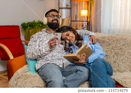 Indian family couple man woman spending calm day at home, reading book listening to music headphones Indian family couple man woman spending calm day at home, reading book listening to music headphones 133714870