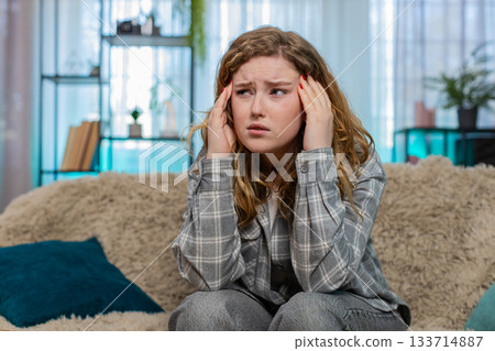 Woman sitting on sofa holding head suffering from strong headache or migraine feeling unwell pain 133714887