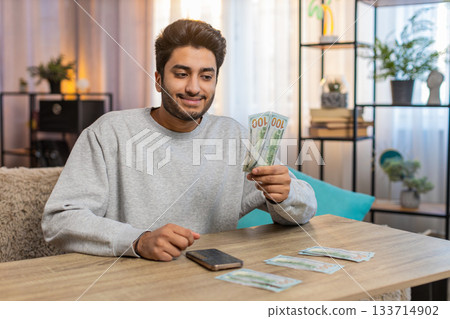 Young Indian man planning expenses, organizing cash in dollars using smartphone calculator at home Young Indian man planning expenses, organizing cash in dollars using smartphone calculator at home 133714902
