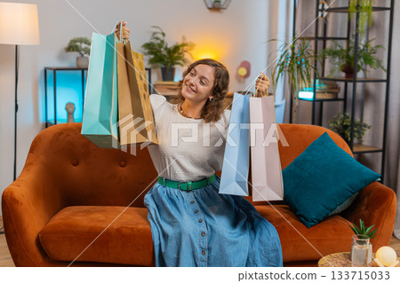 Happy young Caucasian shopaholic woman with shopping bags sitting on sofa in living room at home Happy young Caucasian shopaholic woman with shopping bags sitting on sofa in living room at home 133715033