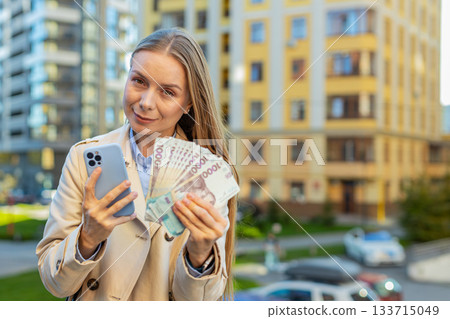 Happy rich mature businesswoman lady using smartphone, holding money hryvnias cash on city street Happy rich mature businesswoman lady using smartphone, holding money hryvnias cash on city street 133715049