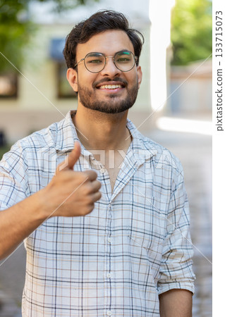 Happy young Indian man looking approvingly at camera showing thumbs up like gesture on city street 133715073