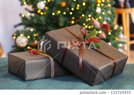 Two Christmas gift boxes decorated with ribbon and red holly berry in front of glowing festive tree Two Christmas gift boxes decorated with ribbon and red holly berry in front of glowing festive tree 133720455