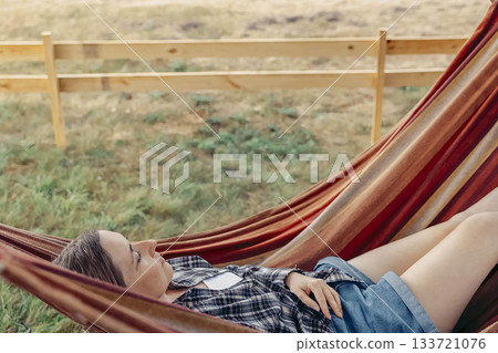 young Caucasian woman with long brown hair relaxes in a colorful hammock under a tree in a rural setting. The scene conveys a sense of tranquility and escape 133721076