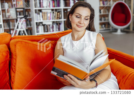 Hispanic woman with short brown hair sits on an orange couch in a library, reading a book. Bookshelves filled with books are visible in the background. Hispanic woman with short brown hair sits on an orange couch in a library, reading a book. Bookshelves filled with books are visible in the background. 133721080