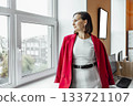 A young Hispanic woman with short dark hair stands by a window in an office. She wears a red blazer over a white dress, looking thoughtfully outside. 133721107
