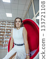 A young Caucasian woman with brown hair sits in a modern red chair in an office library. Bookshelves filled with books are visible in the background. 133721108